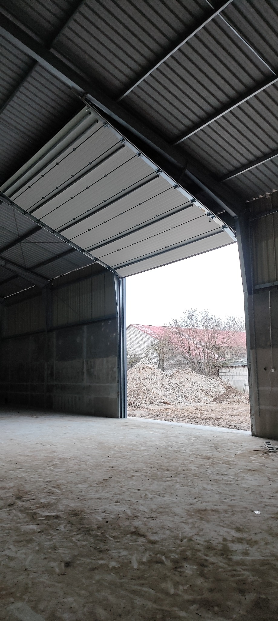 Otwarta brama segmentowa w hali przemysłowej, widok z wnętrza na zewnątrz, gdzie widać gruz i budynki. Szara kolorystyka, surowy klimat.
