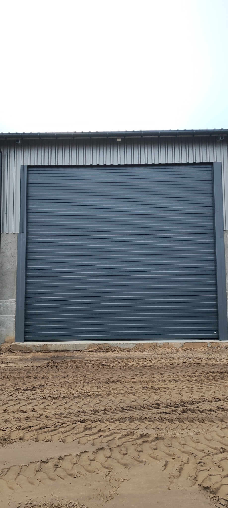 Szara, segmentowa brama garażowa z poziomymi przetłoczeniami, zamontowana w budynku o metalowej konstrukcji. Na dole widoczne ślady opon na ziemi.