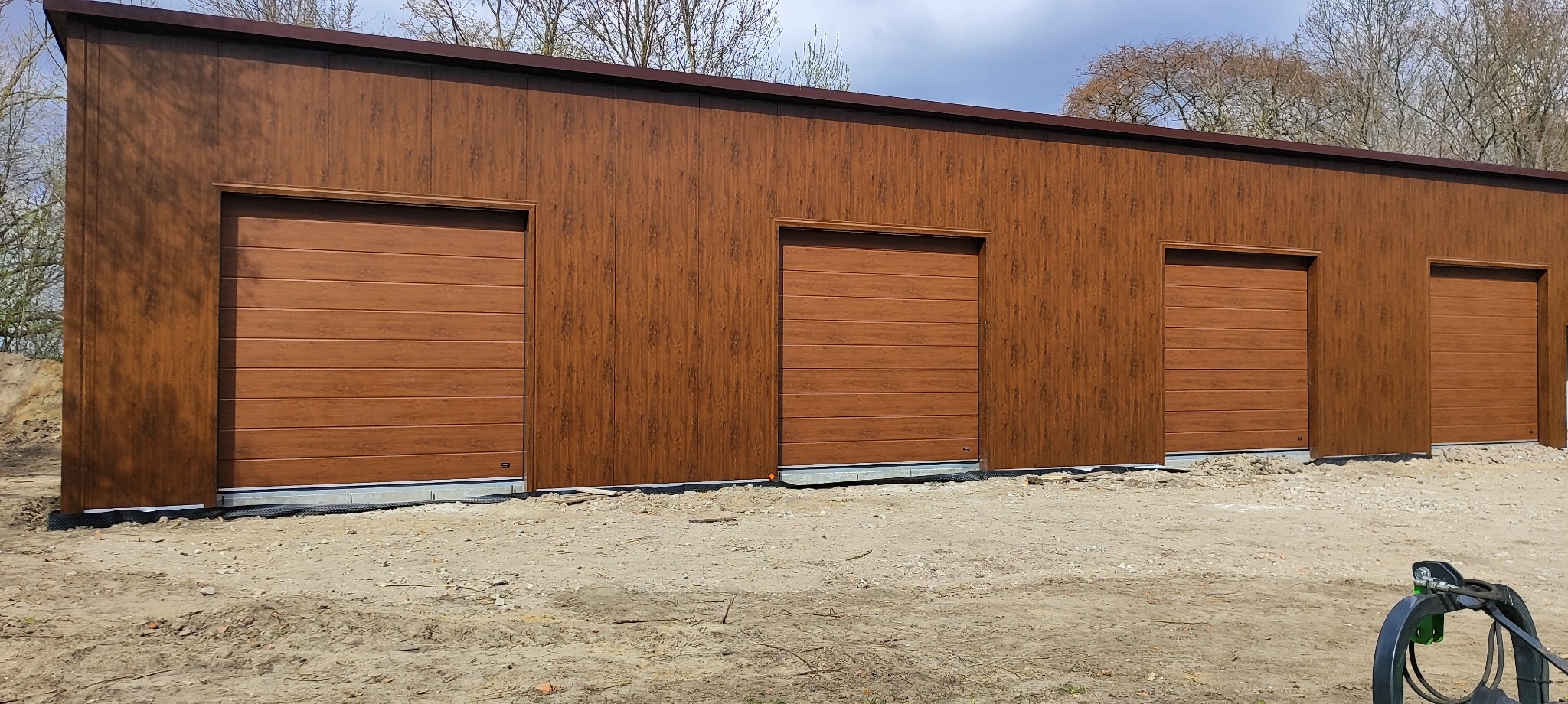 Brązowy budynek z czterema segmentowymi bramami garażowymi o drewnopodobnej fakturze. Widok z zewnątrz, w tle drzewa.