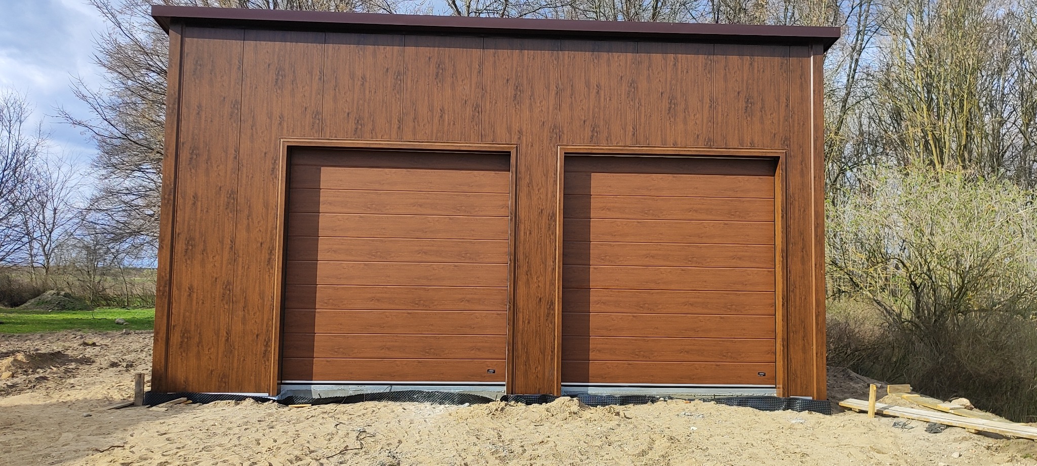 Brązowy garaż z dwiema segmentowymi bramami. Elewacja i bramy w odcieniach drewna. Widoczne elementy konstrukcyjne i piasek wokół, sugerujące świeży montaż.