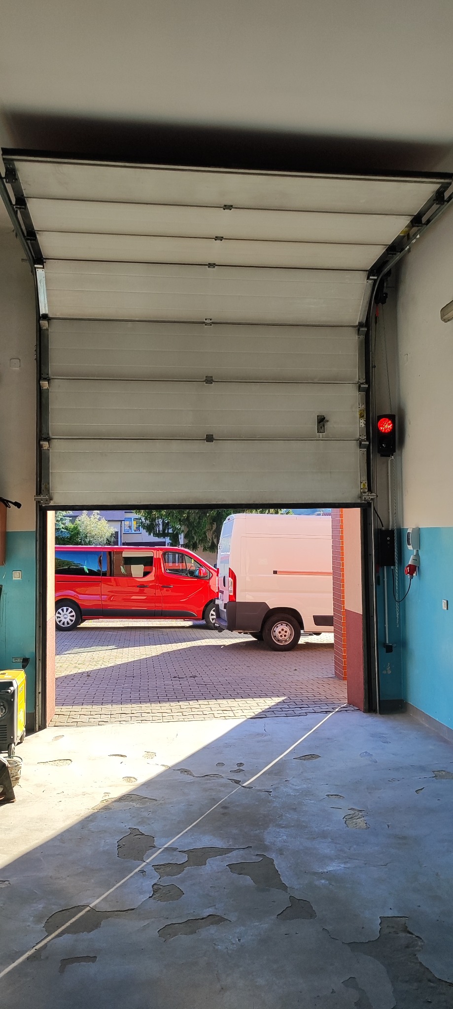 Otwarta segmentowa brama garażowa z sygnalizacją świetlną, widok z wnętrza garażu na podjazd z dwoma samochodami dostawczymi w słoneczny dzień.