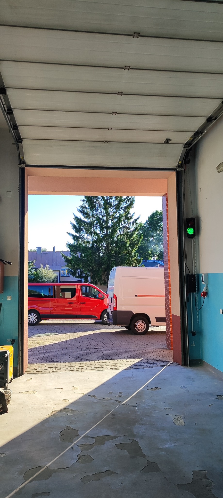 Otwarta brama garażowa z widokiem na podjazd z dwoma samochodami. Widoczny sygnalizator świetlny i mechanizm bramy. Jasne, naturalne światło dzienne.