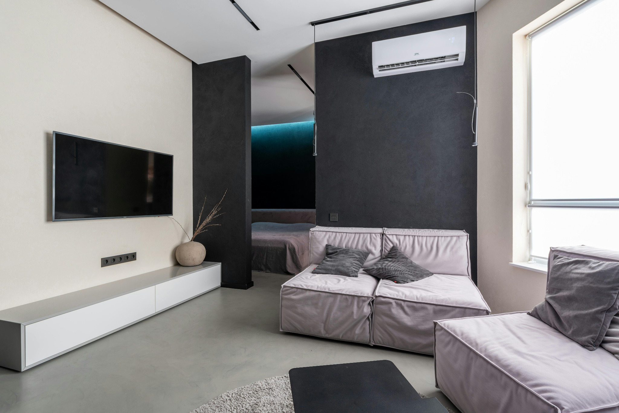 Nowoczesne wnętrze salonu z klimatyzatorem na ciemnej ścianie, minimalistyczny design, jasne sofy i telewizor na tle beżowej ściany. Widok na sypialnię w tle.