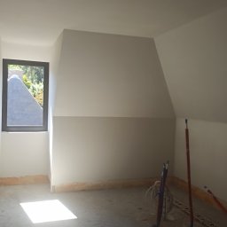 M.R TYNKI - Pokój na poddaszu po nałożeniu gładzi, widoczne instalacje hydrauliczne i okno z widokiem na zewnątrz. Świeżo wykończone ściany i sufit.