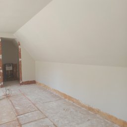 M.R TYNKI - Poddasze po pracach wykończeniowych: białe ściany i sufit, przygotowane podłoże. Widoczne ślady po remoncie i fragment instalacji w przejściu do innego pomieszczenia.