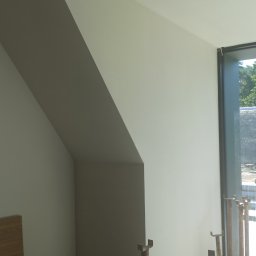 M.R TYNKI - Gładka, jasna ściana po pracach wykończeniowych, widoczne podpory budowlane i fragment okna z widokiem na zieleń. Ściana w kształcie skosu.