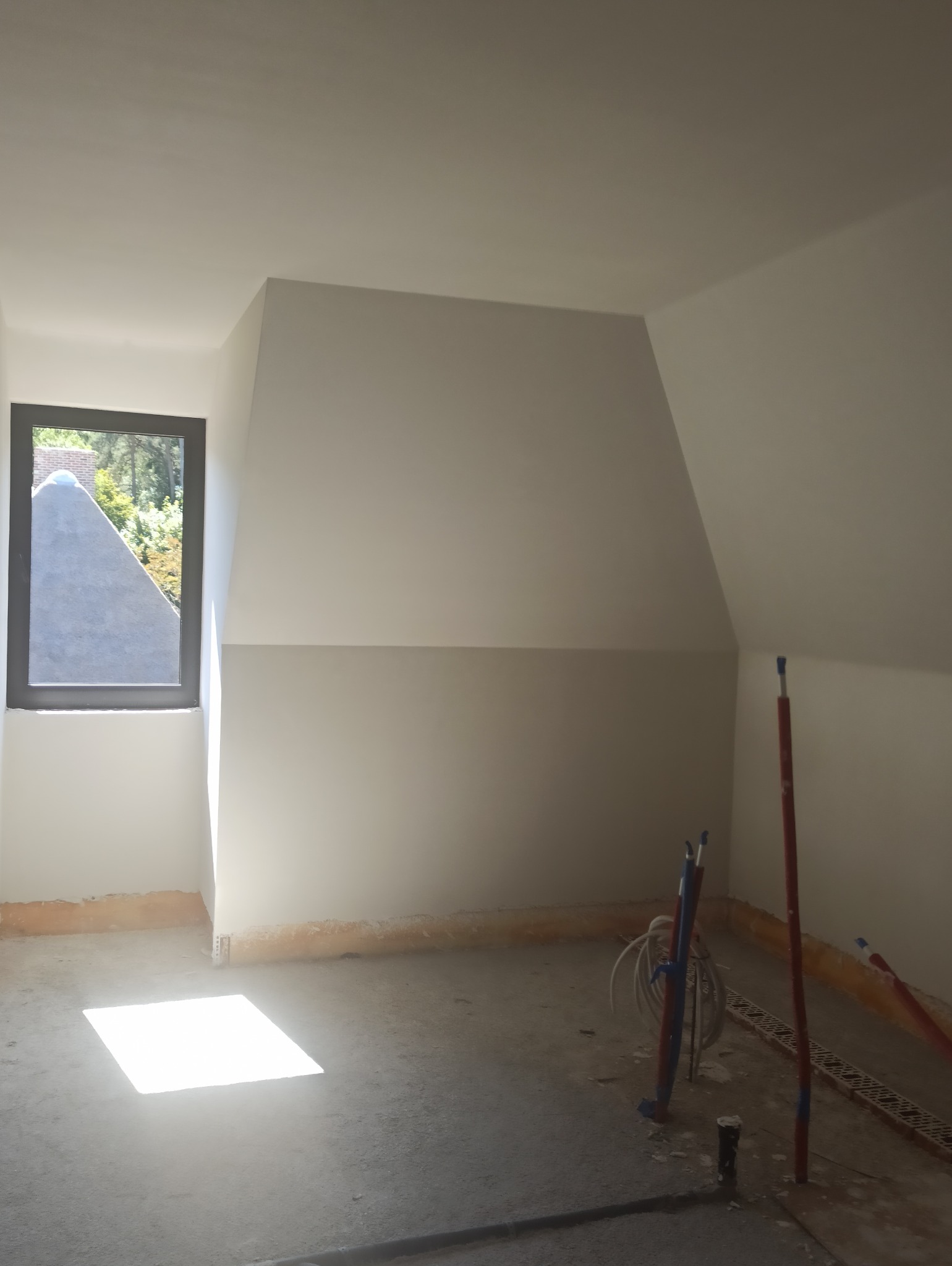 Pokój na poddaszu po nałożeniu gładzi, widoczne instalacje hydrauliczne i okno z widokiem na zewnątrz. Świeżo wykończone ściany i sufit.