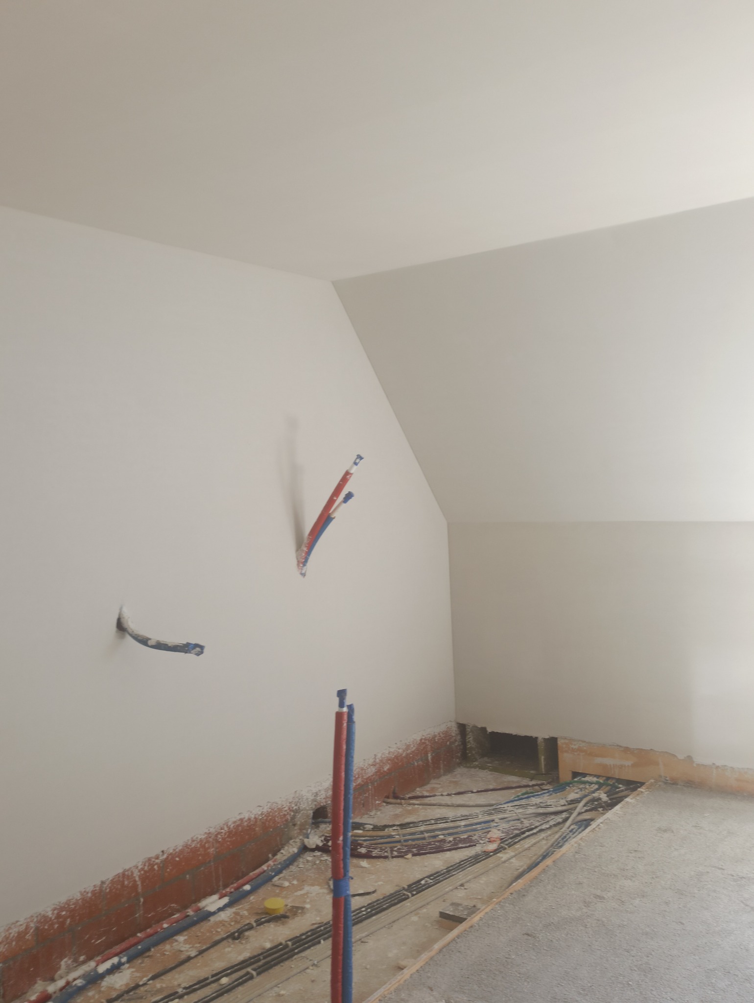 Ściana poddasza z instalacją wodną i elektryczną przed położeniem tynku. Widoczne kable i rury w ścianie oraz podłodze. Surowy stan wykończenia.