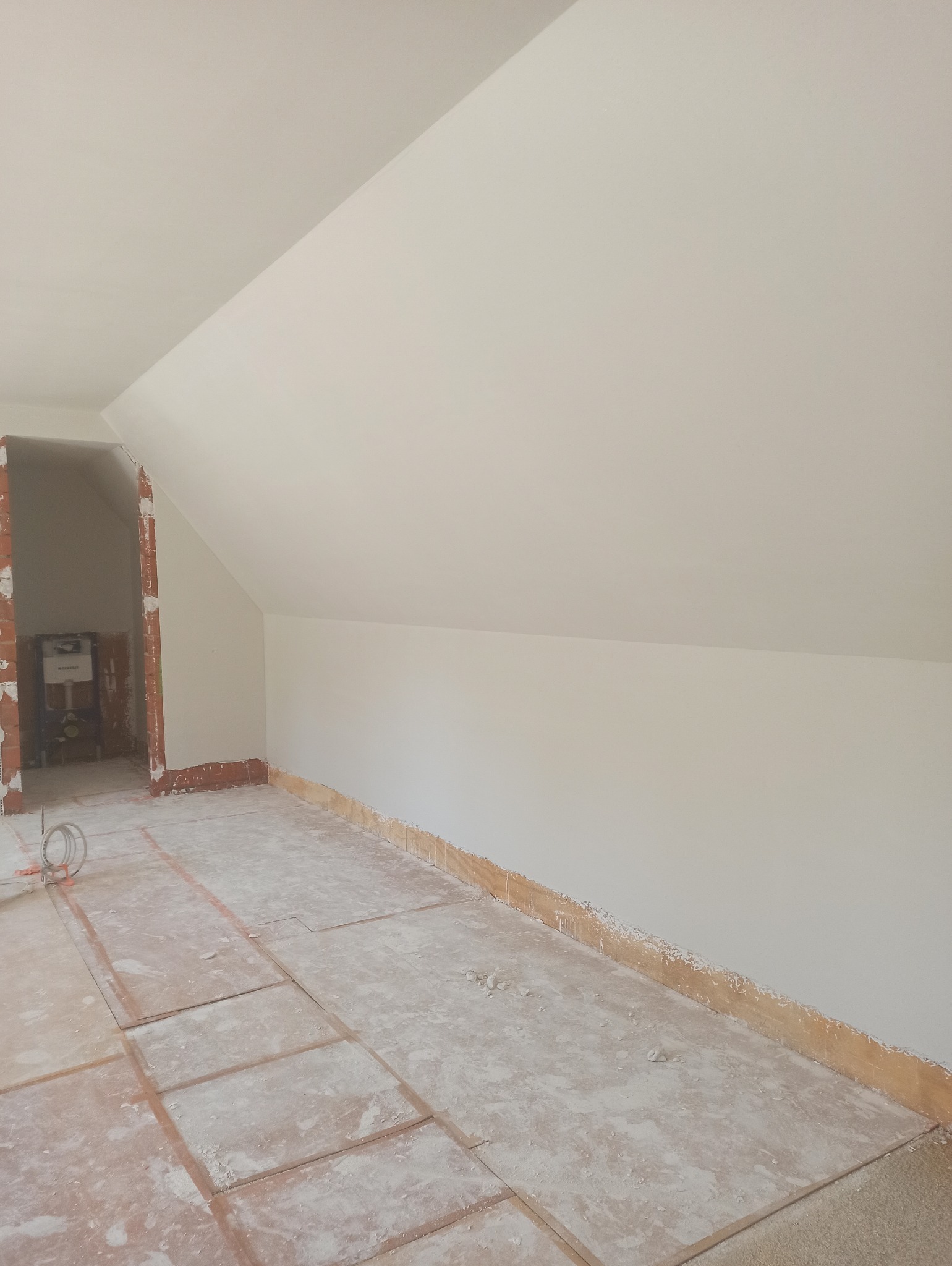 Poddasze po pracach wykończeniowych: białe ściany i sufit, przygotowane podłoże. Widoczne ślady po remoncie i fragment instalacji w przejściu do innego pomieszczenia.