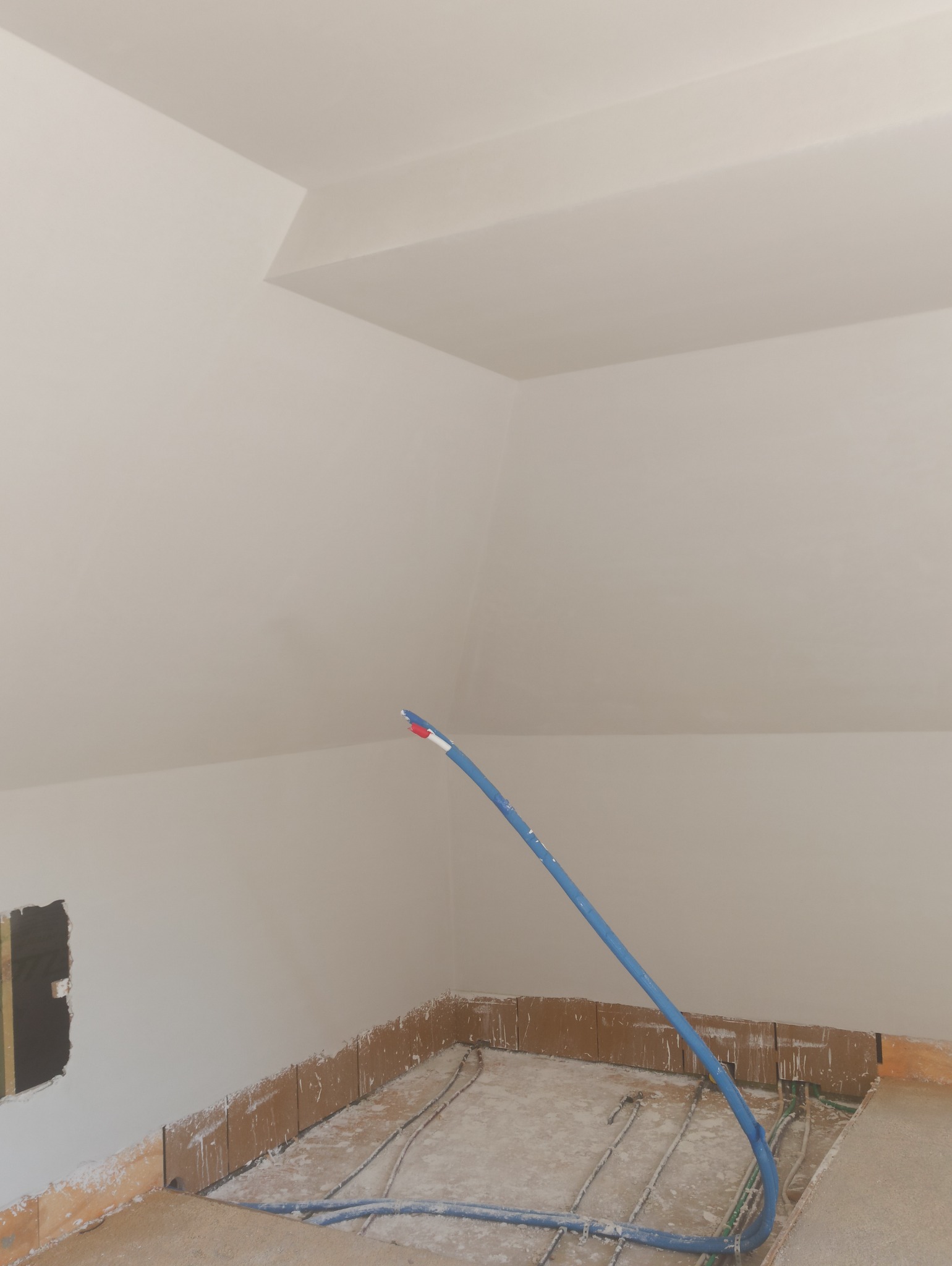Świeżo otynkowane poddasze ze ścianami w kolorze złamanej bieli i instalacją ogrzewania podłogowego z niebieskimi rurami. Widoczny fragment ściany z otworem.