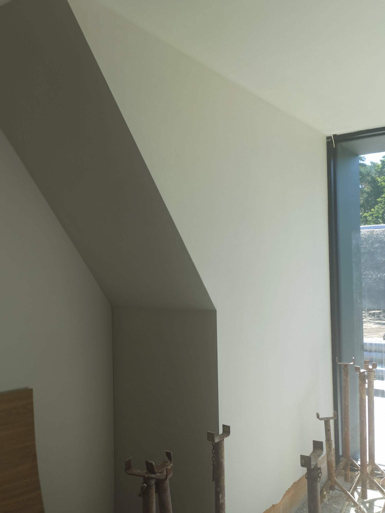 Gładka, jasna ściana po pracach wykończeniowych, widoczne podpory budowlane i fragment okna z widokiem na zieleń. Ściana w kształcie skosu.