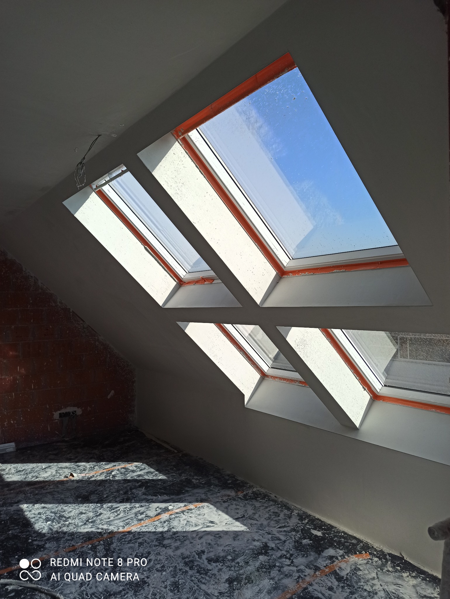 Poddasze po nałożeniu gładzi gipsowej, widoczne okna dachowe i fragment ściany z cegły. Podłoga zabezpieczona folią malarską. Jasne wnętrze w trakcie wykańczania.