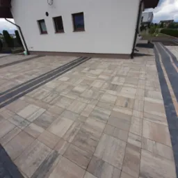 Ułożona kostka brukowa w Kościelnej Wsi, tworząca wzór z ciemniejszych pasów, przylegająca do białego budynku z brązowymi oknami.