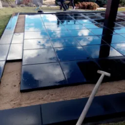 Montaż błyszczących, czarnych płyt tarasowych na piaskowym podłożu, z odbiciem nieba i chmur na powierzchni, widoczna łopata i fragment konstrukcji z drewna.