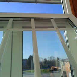 CLEAN PRO SERVICE IVANNA BATENKO - Białe ramy okienne z widokiem na błękitne niebo i zieleń drzew. Czyste szyby odbijają krajobraz. Perspektywa z wnętrza budynku, słoneczny dzień.