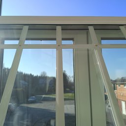 CLEAN PRO SERVICE IVANNA BATENKO - Białe okno z widokiem na zewnątrz, gdzie widać domy i drzewa pod błękitnym niebem. Okno podzielone na kilka sekcji, z wyraźnymi refleksami światła na szybach.