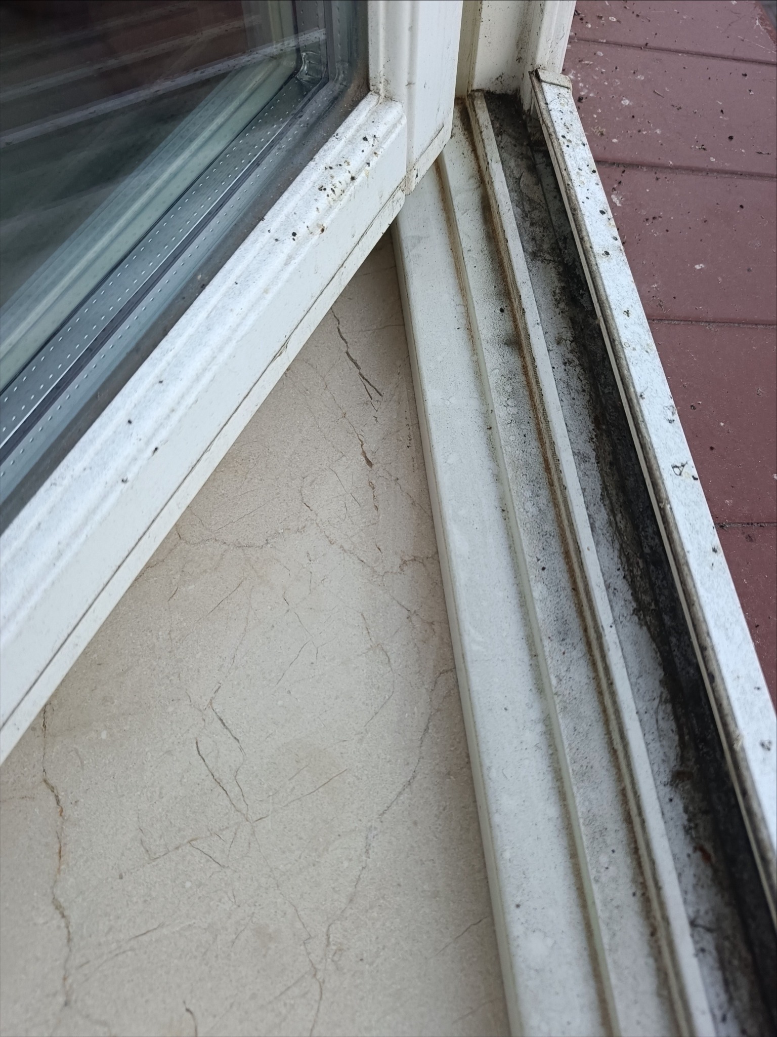 Brudne, białe ramy okienne z widocznymi zabrudzeniami i pleśnią w narożnikach i na uszczelkach. Widoczny fragment szyby i parapetu z beżowego marmuru.