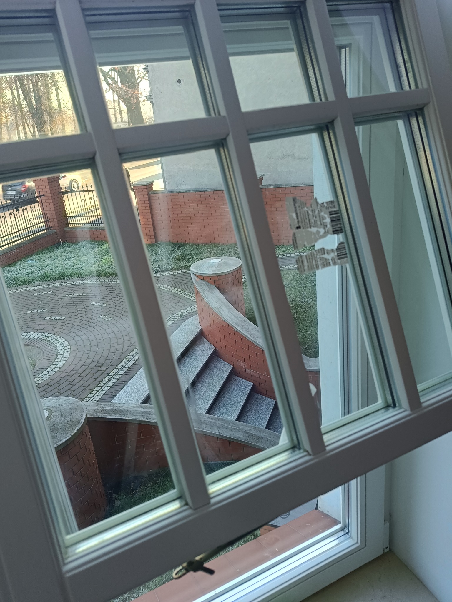 Widok z okna z białymi szprosami na oszroniony ogród z ceglanymi elementami architektury. Otwarta rama okienna, widoczne ślady po naklejce na szybie.