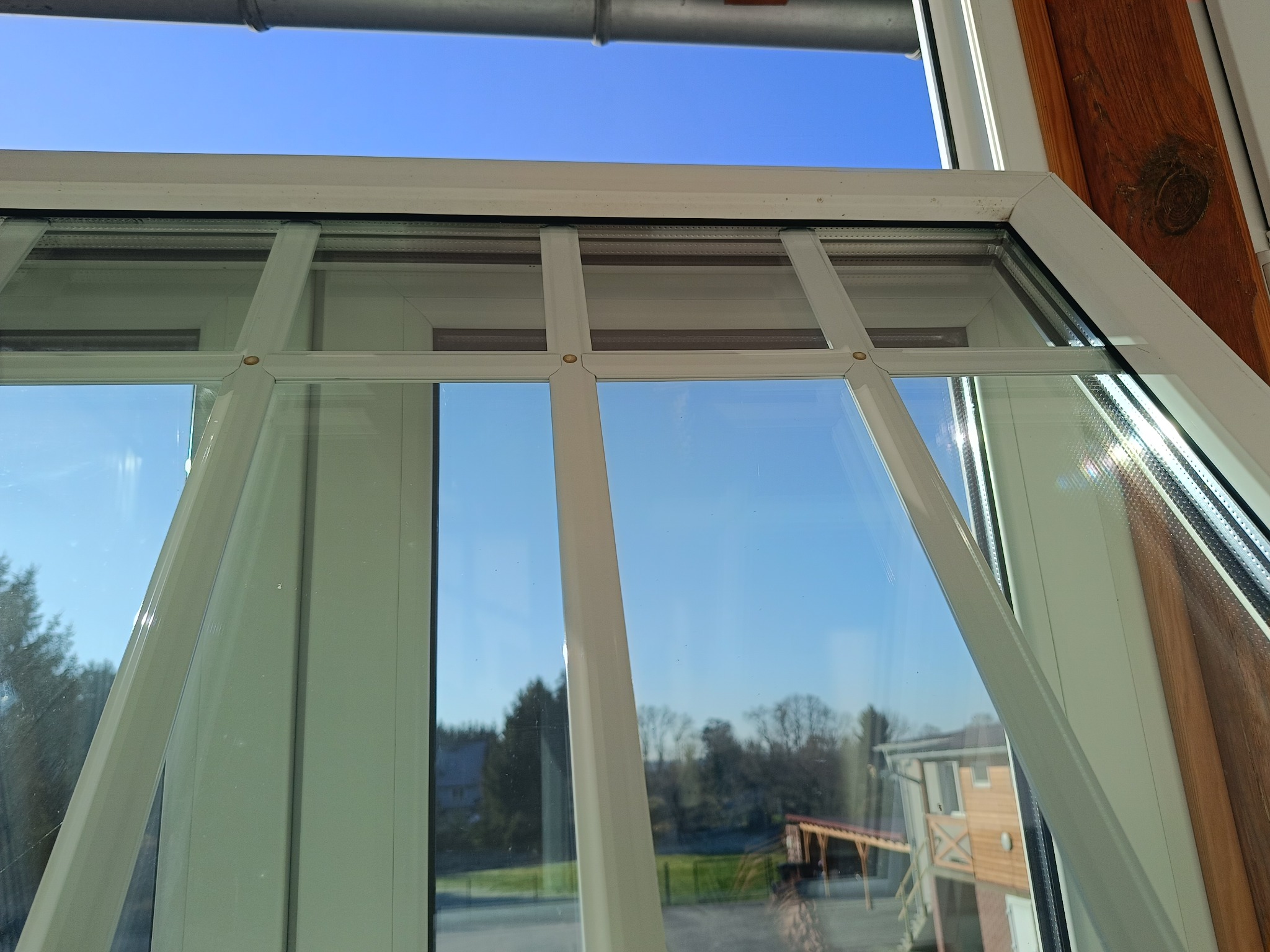 Białe ramy okienne z widokiem na błękitne niebo i zieleń drzew. Czyste szyby odbijają krajobraz. Perspektywa z wnętrza budynku, słoneczny dzień.