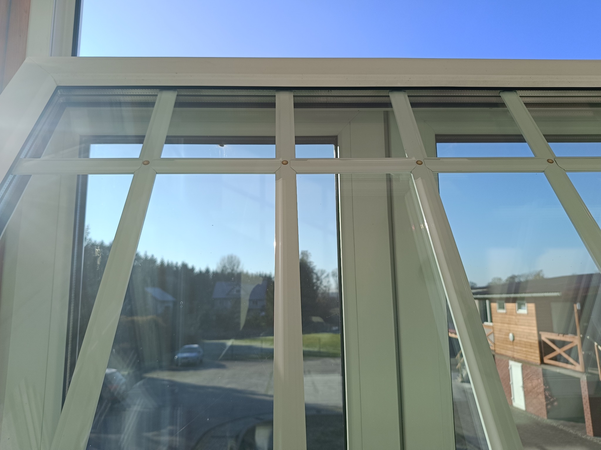 Białe okno z widokiem na zewnątrz, gdzie widać domy i drzewa pod błękitnym niebem. Okno podzielone na kilka sekcji, z wyraźnymi refleksami światła na szybach.