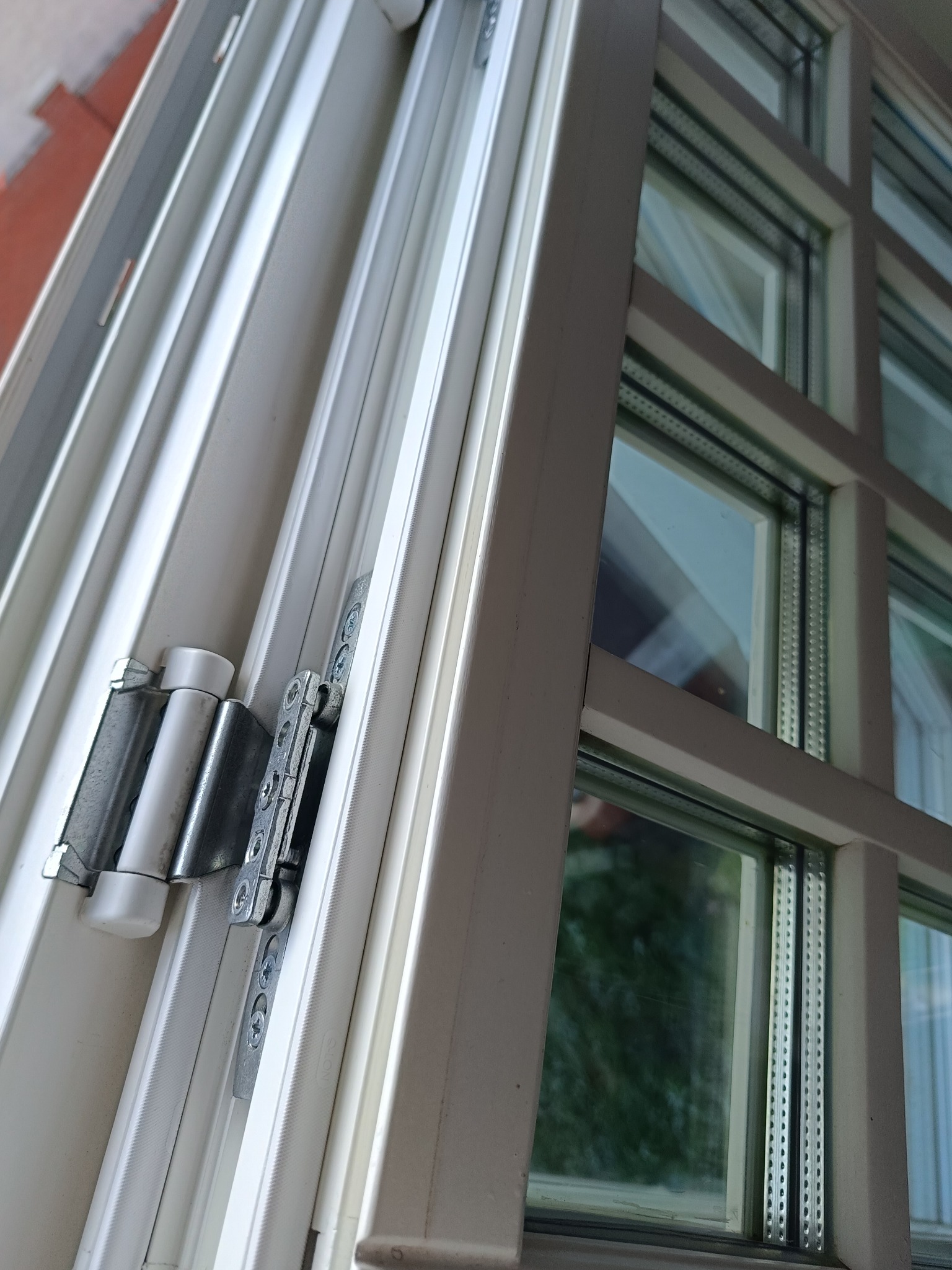 Białe okno PCV z widocznym mechanizmem otwierania, uchylone. Widoczne śruby i detale ramy okiennej. Ujęcie z bliska, z boku.