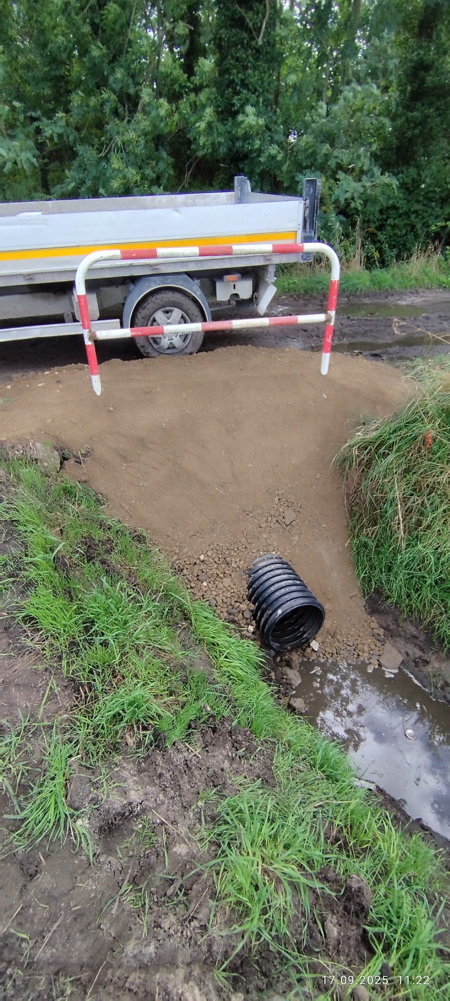 Częściowy widok wykopu pod rurę drenażową i ciężarówki w Rawiczu. Rura leży obok wykopu, w tle zieleń i drzewa. Prace ziemne w toku.