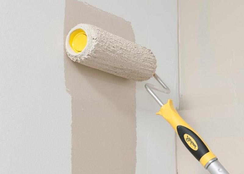 Wałek z żółtą rączką maluje ścianę na beżowo. Widoczny fragment pomalowanej powierzchni i świeża warstwa farby. Detal z bliska, oświetlenie dzienne.