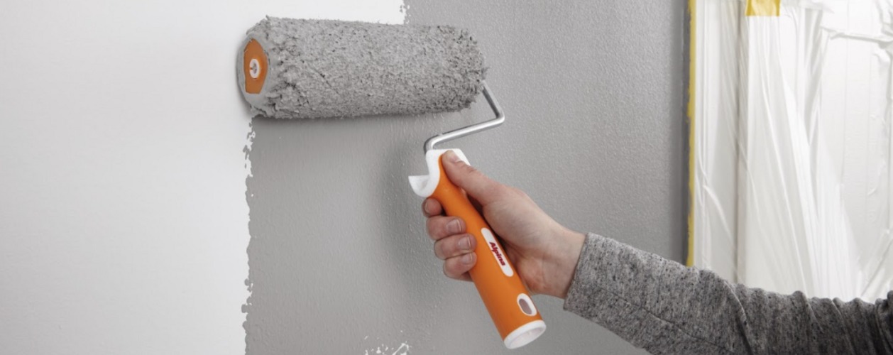 Wałek malarski w akcji, pokrywający szarą farbą ścianę. Częściowo pomalowana ściana na biało, zabezpieczone folią okno. Detal z bliska, oświetlenie dzienne.