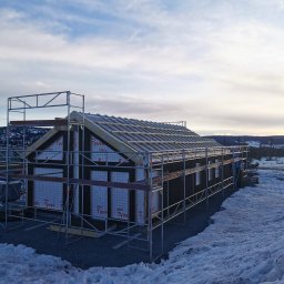 ENFORM Rafał Zulewski - Wznoszenie domu szkieletowego w Sanoku. Widoczne rusztowania, konstrukcja dachu i ściany z izolacją. Zimowy krajobraz w tle, pochmurne niebo.