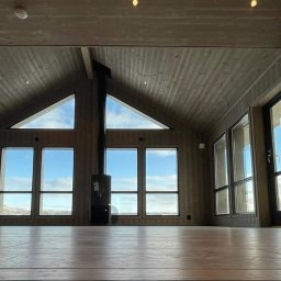 ENFORM Rafał Zulewski - Wnętrze domu z drewnianą boazerią, dużymi oknami z widokiem na krajobraz i kominkiem. Charakterystyczne trójkątne okna pod sufitem.