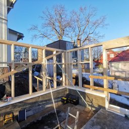 ENFORM Rafał Zulewski - Wznoszenie drewnianej konstrukcji szkieletowej na fundamencie. Widoczne narzędzia, elementy izolacji i otaczająca zabudowa. Słoneczny dzień.