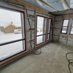 ENFORM Rafał Zulewski - Wnętrze domu w trakcie budowy, widoczne okna z czarnymi ramami, izolacja ścian z folią i drewniane elementy konstrukcyjne. Na zewnątrz śnieg i budynek.