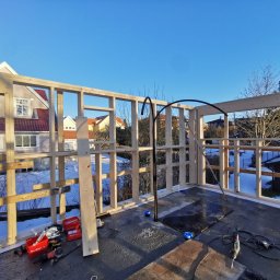 ENFORM Rafał Zulewski - Wznoszenie drewnianej konstrukcji szkieletowej na dachu budynku w słoneczny dzień. Widoczne narzędzia i materiały budowlane. W tle zabudowa jednorodzinna.