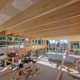 ENFORM Rafał Zulewski - Wnętrze domu w trakcie budowy szkieletowej. Widoczne drewniane belki stropowe, ściany w konstrukcji szkieletowej, narzędzia budowlane i materiały. Jasne, naturalne światło.