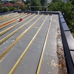 ENFORM Rafał Zulewski - Konstrukcja dachu płaskiego z drewnianymi belkami na szarej izolacji, widok z góry, w tle zieleń drzew i zabudowa miejska w słoneczny dzień.