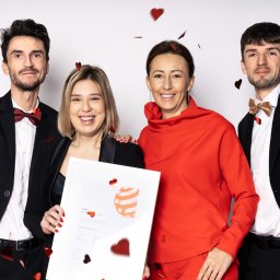 MAKE THE FLOW - Cztery uśmiechnięte osoby w eleganckich strojach pozują z nagrodą Red Dot Award na białym tle, z konfetti w kształcie serc. Kobieta trzyma certyfikat.