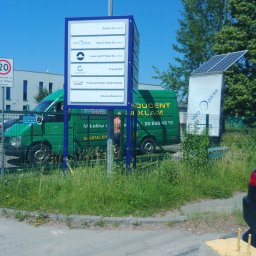 SCAN MOTOPOL SPÓŁKA Z OGRANICZONĄ ODPOWIEDZIALNOŚCIĄ - Tablica informacyjna z logotypami firm, zielony van z reklamą na tle budynku i zieleni. Dodatkowo tablica z panelem solarnym. Słoneczny dzień.