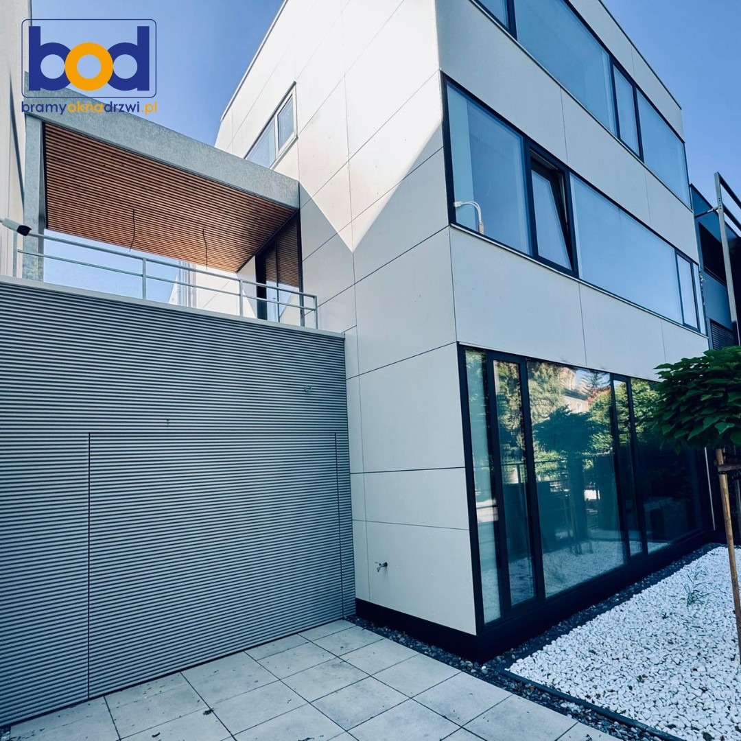 Nowoczesny budynek z szarą bramą garażową z poziomymi panelami, dużymi oknami i balkonem z drewnianym sufitem. Białe kamienie dekoracyjne u podstawy.