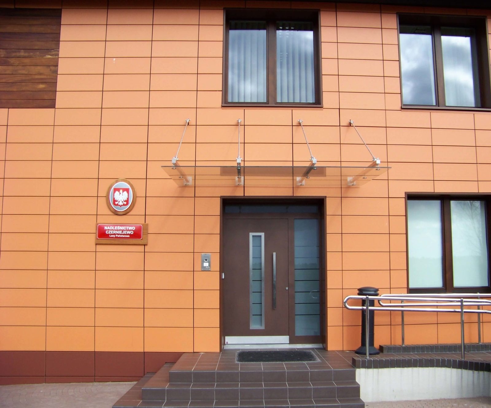 Brązowe drzwi wejściowe z przeszklonym panelem bocznym i daszkiem, na tle pomarańczowej elewacji budynku z oknami. Widoczne schody wejściowe i balustrada.