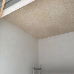 MAM Remonty - Montaż paneli sufitowych z włókna drzewnego w jasnym wnętrzu. Widoczny fragment rusztowania i surowa ściana. Ujęcie z dołu, podkreślające strukturę paneli.