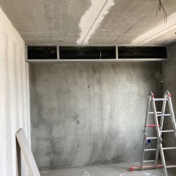 MAM Remonty - Surowy pokój w trakcie wykończenia: szpachlowana ściana, metalowa konstrukcja pod sufitem, drabina i płyty karton-gips. Widoczne kable elektryczne.