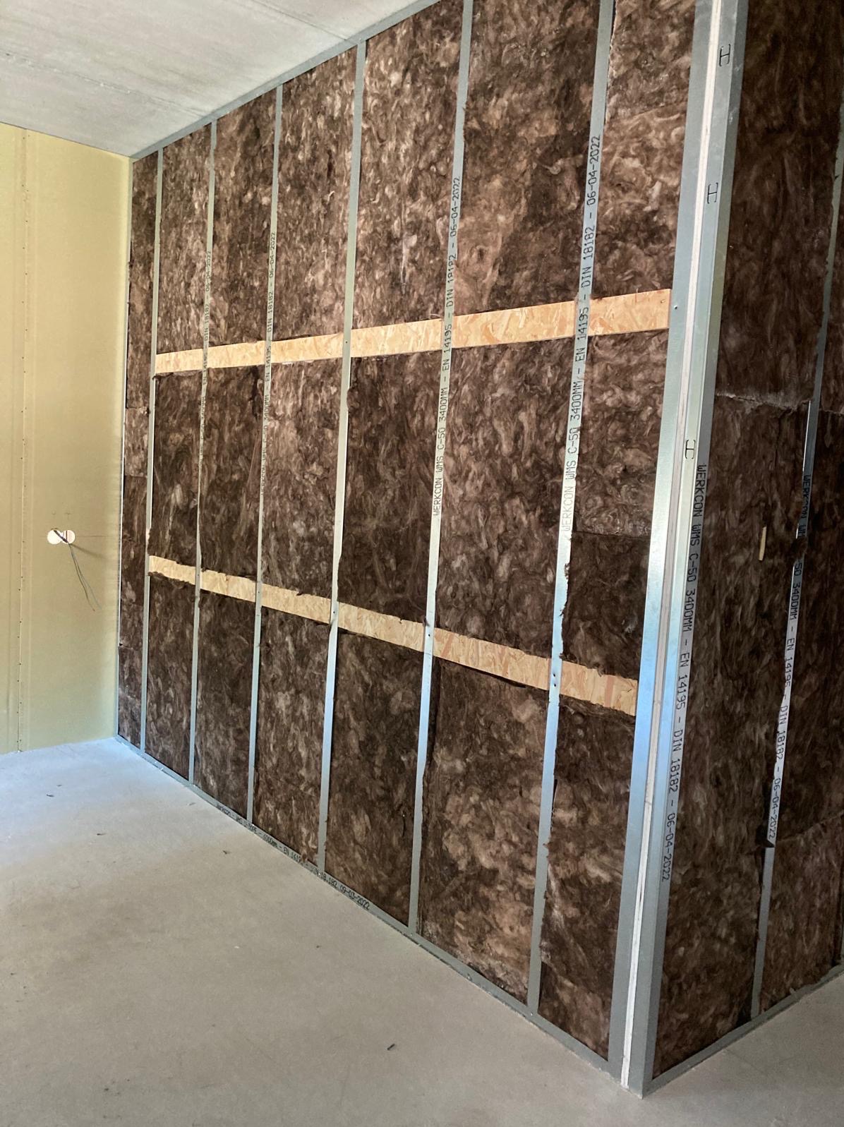 Ściana z izolacją z wełny mineralnej, widoczne metalowe profile i drewniane poprzeczki. Etap prac wykończeniowych w jasnym pomieszczeniu.