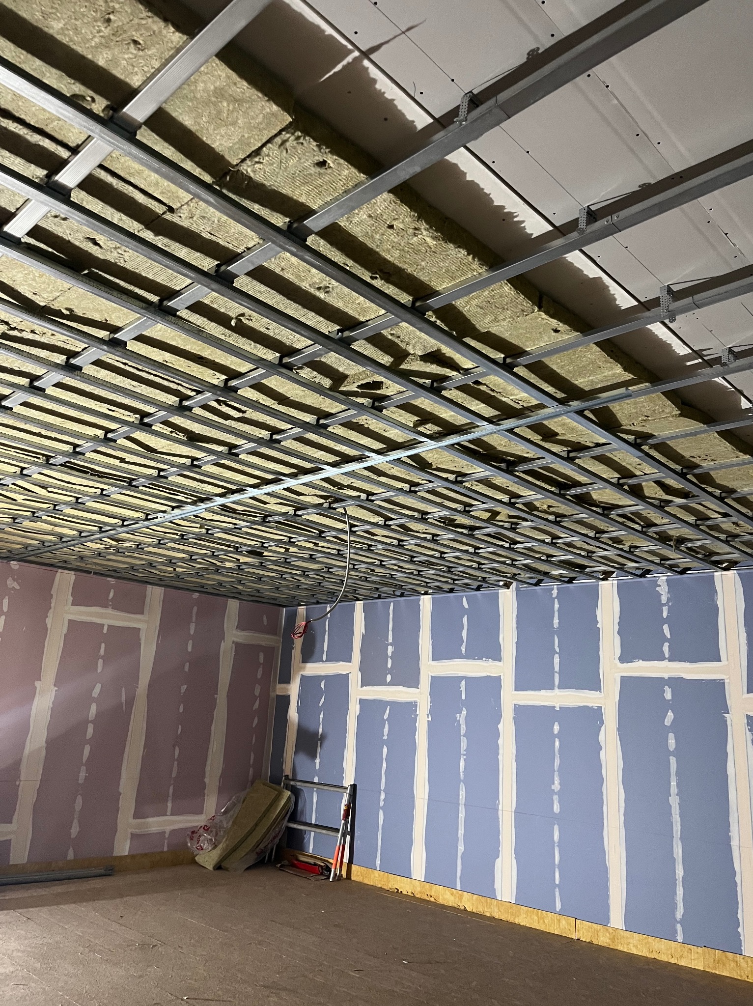 Sufit podwieszany z izolacją z wełny mineralnej i częściowo zamontowanymi płytami. Ściany z płytami w kolorach fioletu i niebieskiego, przygotowane do dalszych prac.
