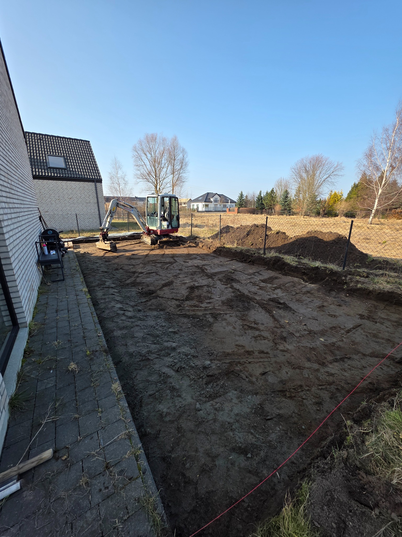 Mini koparka na działce z przygotowanym terenem pod budowę, widoczne ogrodzenie i budynek w tle, słoneczny dzień.