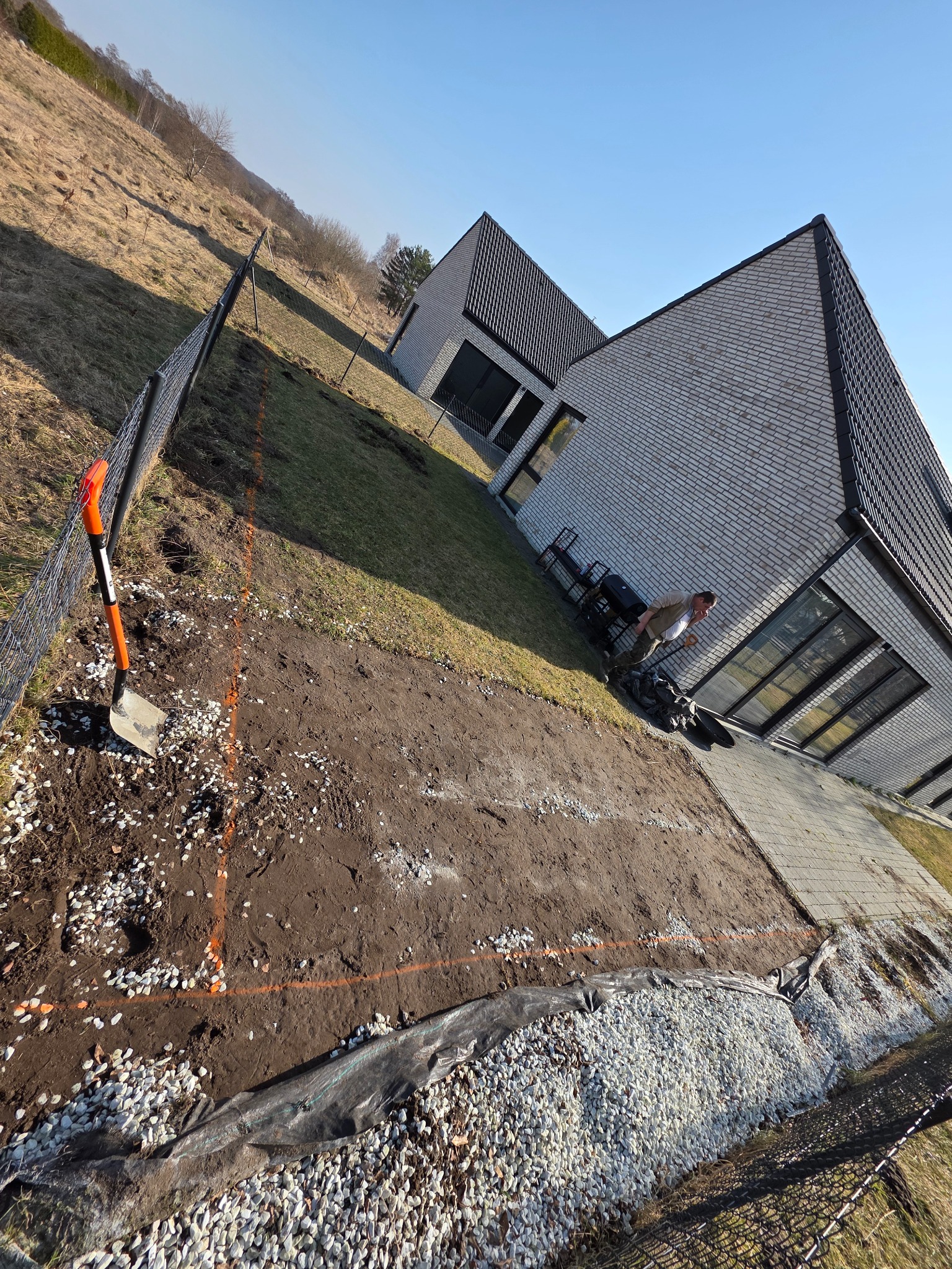 Przygotowanie terenu pod kostkę w Ustce: wyznaczony obszar, niwelacja ziemi, podsypka z kamienia, widoczna łopata i nowoczesny dom w tle. Prace ziemne.