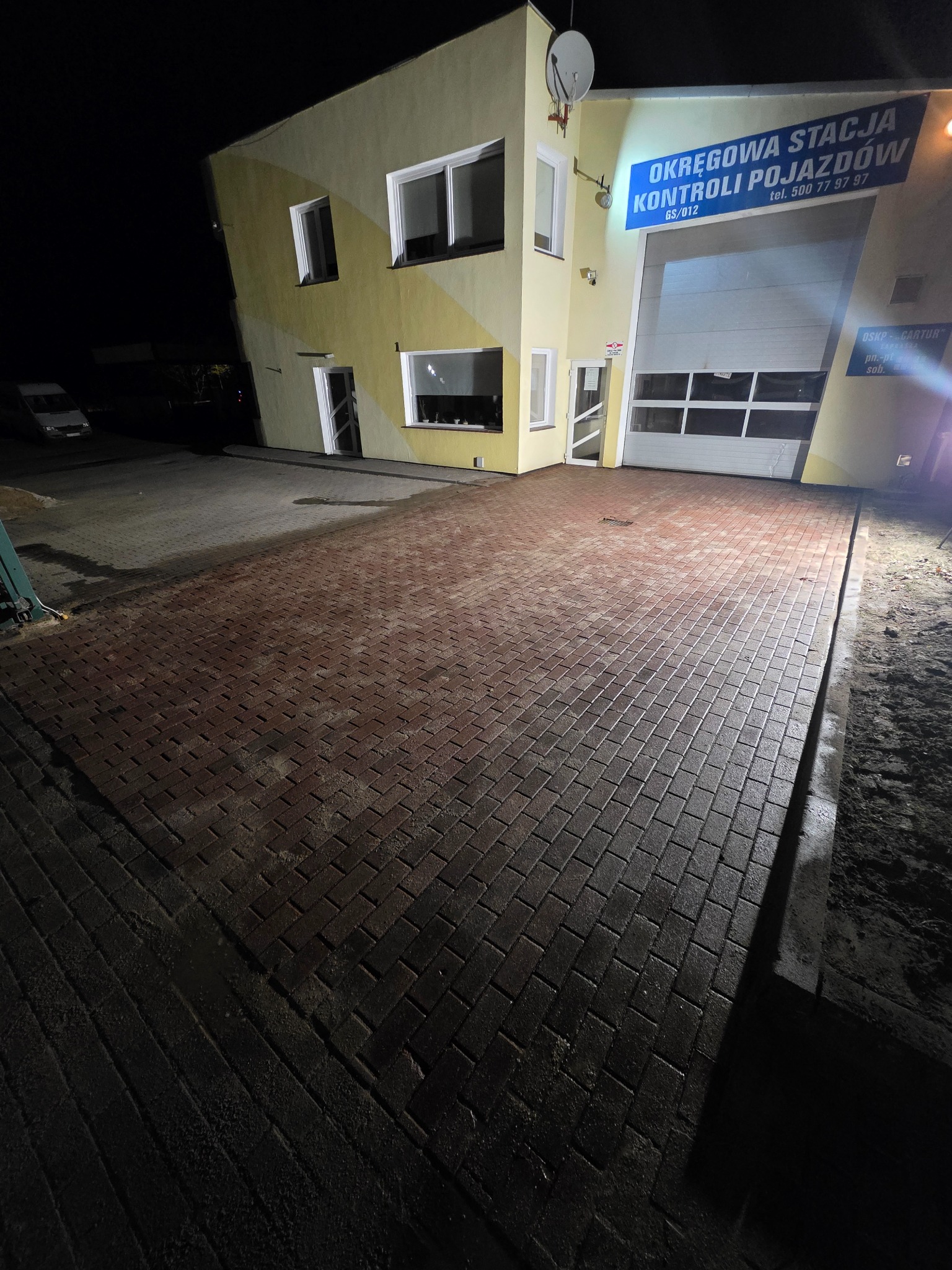 Czerwona kostka brukowa przed budynkiem stacji kontroli pojazdów, oświetlona nocą. Widoczny fragment budynku z bramą garażową i szyldem informacyjnym.