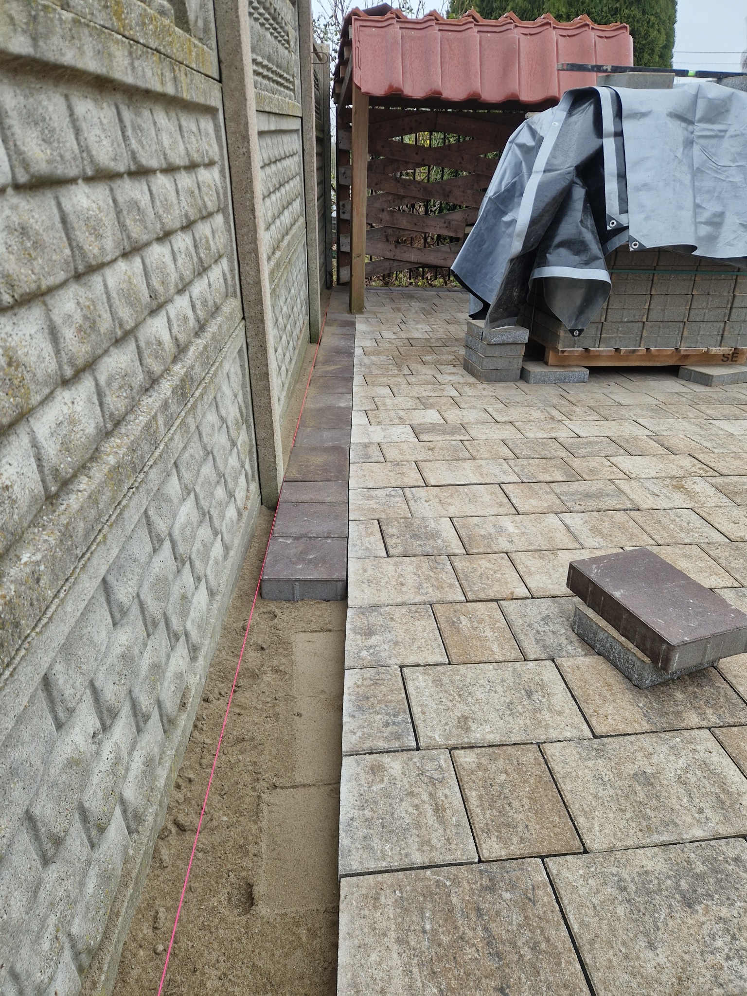 Fragment układanej kostki brukowej wzdłuż betonowego ogrodzenia. Widoczny piasek, sznurek wyznaczający linię i paleta z kostką pod plandeką w tle.