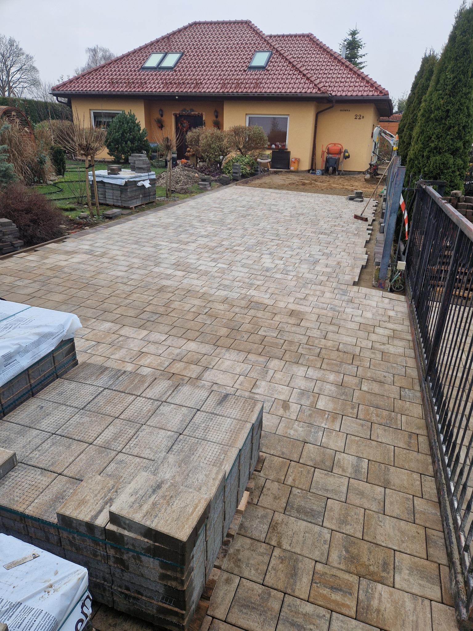 Ułożona kostka brukowa na podjeździe domu jednorodzinnego. Częściowo ułożona nawierzchnia, palety z kostką i mini koparka w tle. Widok z góry.