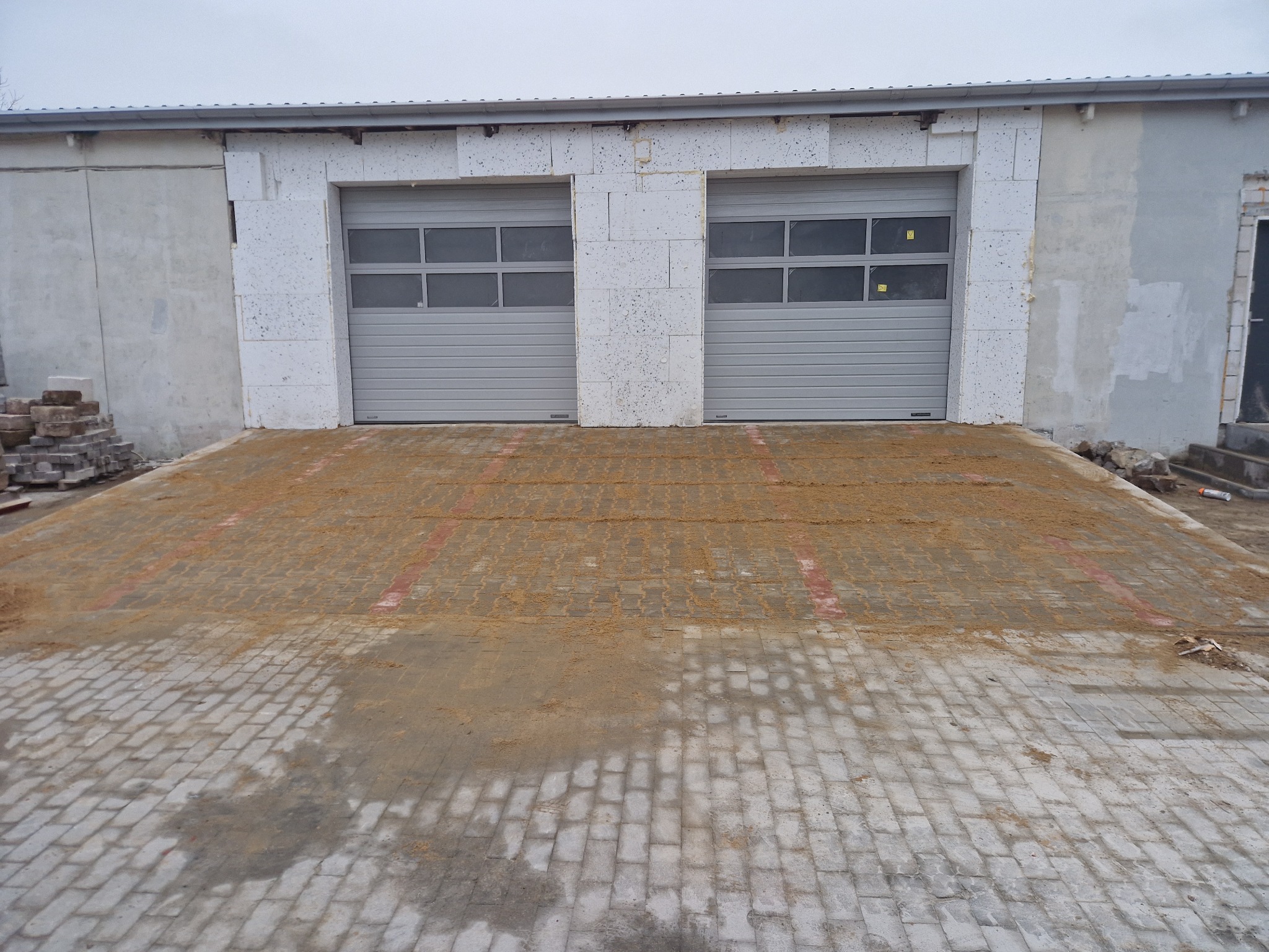 Ułożona kostka brukowa przed budynkiem z dwoma garażami, częściowo zasypana piaskiem. Widoczne niedociągnięcia w ułożeniu. Kostka w odcieniach szarości.