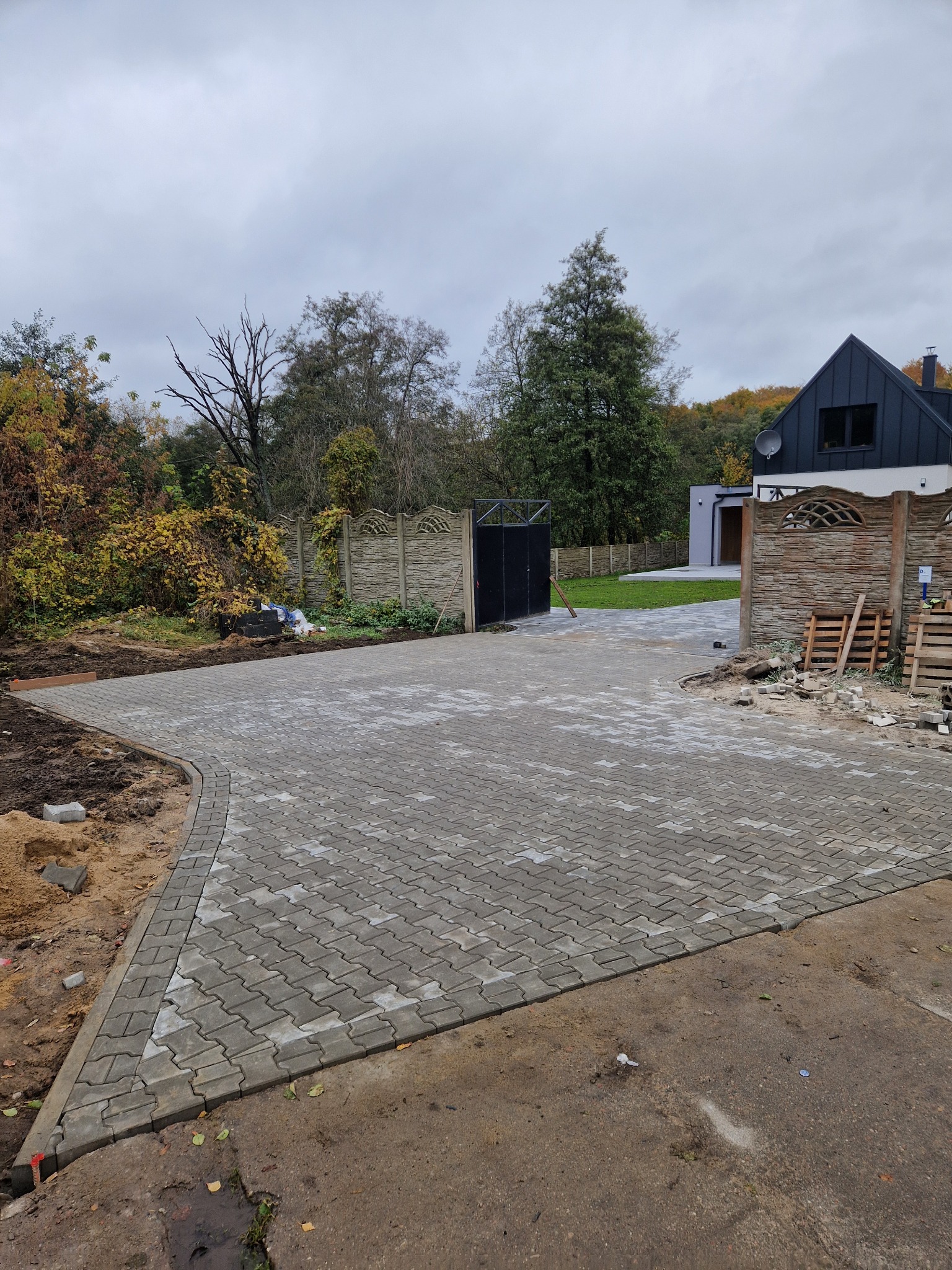 Ułożona szara kostka brukowa na podjeździe domu jednorodzinnego, widok na bramę i ogrodzenie. Częściowo widoczne palety i resztki materiałów budowlanych.